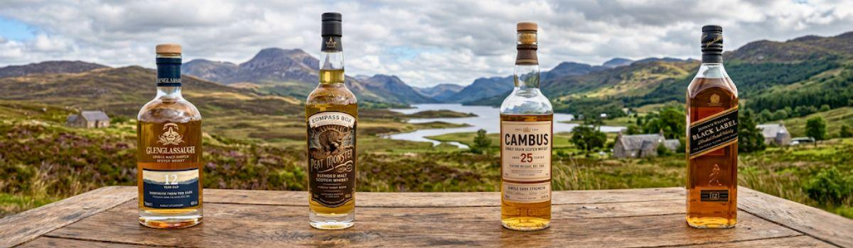 verschiedene Whiskey-Flaschen auf einem Holztisch mit Blick auf die Schottische Landschaft verschiedene Whiskey-Flaschen auf einem Holztisch mit Blick auf die Schottische Landschaft